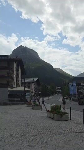 The video pans right across a street scene in a mountain village. The foreground is a cobblestone plaza with two rectangular planters filled with colorful flowers. Several black bollards are visible along the edge of the plaza. In the mid-ground, a paved road curves to the right, with buildings lining its left side. People are walking along the sidewalk on the left side of the road. The buildings are a mix of traditional alpine architecture with wooden balconies and more modern designs. Two blue banners with white lettering hang from a building on the right side of the road. Beyond the village, lush green mountains rise up, with a large, imposing peak dominating the left side of the frame. The sky is cloudy with patches of blue sky visible.