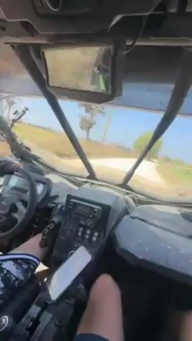 From a POV shot inside an all-terrain vehicle, it drives along a dirt road on a bright day. The road is flanked by fields. The interior of the vehicle is visible, including the steering wheel, dashboard, and rearview mirror. A phone is mounted on the dashboard. The driver wears a black and white patterned shirt and is visible in the bottom left of the frame.