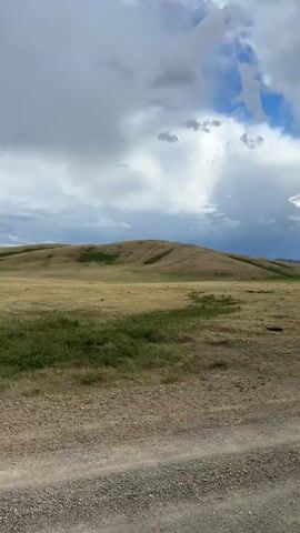 This media file captures a sweeping landscape under a dramatic sky. The foreground is dominated by a rough, gravelly dirt road, suggesting a rural or remote setting. Beyond the road lies a vast expanse of dry, golden-brown grassland, interspersed with patches of greener vegetation. In the mid-ground, a gently sloping hill rises, its crest catching the light.

The sky is a significant element, filled with a mix of heavy, dark clouds and patches of bright blue sky. Rays of sunlight are piercing through the clouds in the distance, creating dramatic shafts of light that illuminate the horizon. This suggests a dynamic weather system, possibly a storm passing or approaching, contributing to a sense of awe and perhaps a touch of melancholy. The time of day appears to be late afternoon or early evening, given the quality of the light and the length of the shadows.

The overall scene evokes a sense of wide-open spaces and the untamed beauty of nature. There are no people or man-made structures immediately visible, emphasizing the natural elements. The location cues of "Bakty, Kazakhstan" place this scene in a vast, potentially arid or semi-arid region. The mood is contemplative, with the grandeur of the landscape and the powerful sky creating a profound sense of isolation and natural wonder. There is no visible text in the media file.