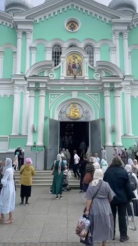 The video shows the entrance to a large, light green church with white trim. The entrance is a grand archway with large, open double doors. Above the archway is a mosaic depicting religious figures. There are steps leading up to the entrance, and a group of people are walking in and out of the church. The people are dressed in casual clothing, and some of the women are wearing headscarves. The ground in front of the church is paved with grey stone. The sky is overcast.