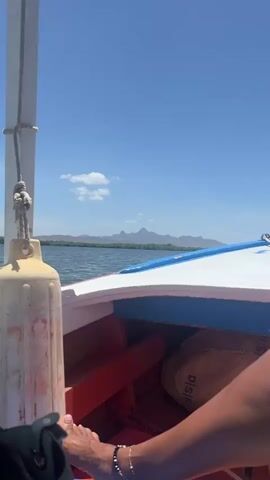 The video shows a boat ride on a sunny day. The camera is positioned inside the boat, with the view extending over the front of the boat towards the water. In the distance, a mountain range is visible. A white and blue painted section of the boat's bow is in view. A dirty, white bumper hangs from a rope tied to a pole to the left. A brown bag sits behind the boat's front. A person's leg and foot are visible at the bottom of the frame, with a black sandal beside their foot and an anklet. The sky is blue with a few white clouds. Birds are flying in the sky. The water is calm, with some ripples.