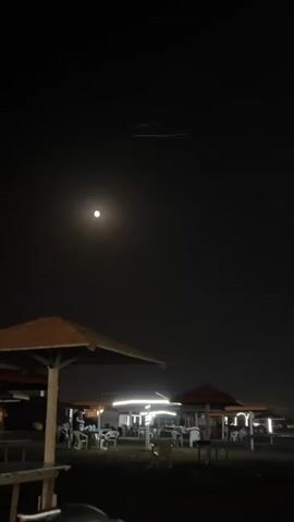A nighttime scene unfolds at a beachside establishment. The sky is dark, illuminated by a bright full moon and a few distant lights. Several open-air structures, likely food stalls or seating areas, are visible, some equipped with simple tables and chairs. String lights hang from some of these structures, casting a warm glow on the ground. A few people are seated at tables, while others are standing or walking around. In the foreground, a person is standing near a wooden structure. Another person walks away from the camera towards the seating areas. The ground appears to be dark sand or dirt. Two faint linear streaks are visible in the upper portion of the sky, possibly trails from an aircraft.