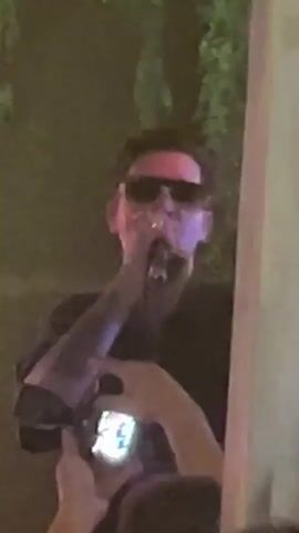 A man wearing sunglasses is singing into a microphone while standing outdoors at night. He is wearing a dark t-shirt and has tattoos on his arms. A person in the foreground is holding up a smartphone, seemingly to record the performance. The background is dark and features greenery, possibly trees or vines, illuminated by green lighting. The overall atmosphere appears to be that of a live music event.