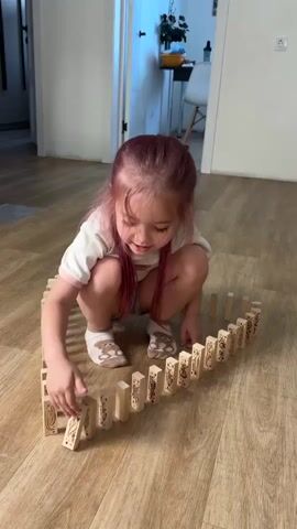 A young girl with pink-dyed hair and a white t-shirt and purple shorts is playing with dominoes on a wooden floor. The dominoes are arranged in a circular or semi-circular pattern. The girl is crouching down and appears to be setting up the dominoes or about to knock them down. The scene is indoors, with a doorway visible in the background. The lighting suggests it could be daytime. The girl seems focused on her activity. There is no visible text in the video. The setting appears to be a residential interior, possibly in Russia given the location context.