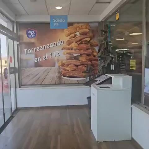 The video shows the interior of a casual dining establishment, possibly a roadside rest stop or a restaurant with a rustic theme. The lighting is bright and fluorescent. The flooring is light-colored wood laminate. To the left, there is a large advertisement on a window or wall featuring a stack of what appears to be fried pork rinds or similar food items, with text that reads "Torrezneando en el 112". Above this is a blue sign with "Salida Exit" written in white. In the foreground, a white reception or service counter is visible, with a small black waste bin next to it. To the right of the counter, a decorative arrangement of tall bamboo stalks in a dark planter stands. Beyond this, the scene opens up to the main dining area. A bar or counter runs along the back wall, with shelves stocked with glasses and bottles. Several televisions are mounted above the bar. The dining area is populated by patrons, with some standing near the bar and others seated at tables. Several decorative barrels, labeled "PATA NEGRA", are placed around the dining area, adding to the rustic aesthetic. The overall impression is of a busy, functional establishment.
