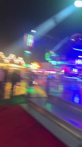 The video shows a blurred view of a fairground at night. Bright, colorful lights from various rides, including a ferris wheel and other attractions, create a dynamic backdrop. People are walking through the fairground, their figures appearing as blurred silhouettes. The camera movement is unsteady, adding to the chaotic atmosphere of the fair.