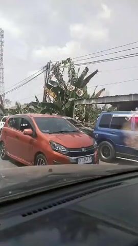 The video shows a street scene with several cars parked on the side of the road. A tall communication tower is visible in the background, along with some trees and buildings. The weather appears to be clear and sunny. The camera is inside a vehicle, looking out at the street. The cars include a white SUV, a black car, a red car, a maroon SUV, a blue van, and a silver pickup truck. Some of the cars are parked close together, suggesting a potentially crowded area. The overall impression is of a typical day on a street in a populated area.