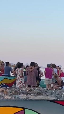A group of people are gathered on a beach. A man wearing a blue shirt and jeans is sitting on a colorful boat. Several women are standing and talking. One woman is wearing a floral dress. Another woman is wearing a pink top and shorts. There is a table with bottles and containers. In the background, there is water and a distant shore with buildings and palm trees. The sky is clear and appears to be during sunset or sunrise.