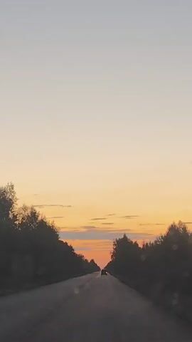 The video shows a view from inside a vehicle driving on a paved road at dusk. The sun is setting on the horizon, casting a warm orange glow across the sky, with soft clouds scattered throughout. Dense trees line both sides of the road, creating a tunnel effect. A single car is visible in the distance, driving in the same direction. The road surface appears to be asphalt.