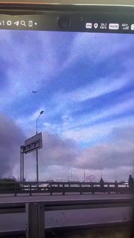 The video captures a wide shot of an outdoor scene. The sky is a prominent feature, with blue patches and scattered clouds. In the upper left of the frame, a small dark object, possibly an aircraft, is visible against the sky. A tall lamppost with a large sign attached stands on the left side. A bridge or elevated roadway stretches horizontally across the mid-ground, with a railing. Several figures, appearing as silhouettes, are visible standing on the bridge. In the right mid-ground, a large plume of dark smoke rises into the sky, indicating a fire or explosion. The lower part of the frame shows grassy terrain and a concrete barrier. The video seems to be recorded from a stationary, possibly elevated, position, looking towards the bridge and the smoky area.