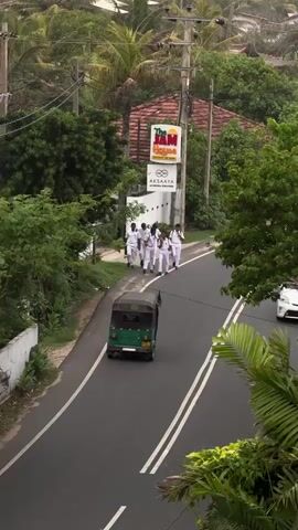 The media depicts a street scene in Ahangama, Sri Lanka. In the foreground, a paved road with double white lines runs diagonally from bottom left to top right. On the left side of the road, a white concrete wall borders a vegetated area. On the right side, lush green foliage, including palm trees, frames the road.

Several subjects are visible. A group of approximately seven individuals, all wearing white uniforms, are walking along the side of the road. They appear to be moving away from the camera. A green tuk-tuk is driving on the road, passing the group of pedestrians. Further down the road, a white car is parked or moving slowly. A person on a motorcycle, wearing a pink shirt and dark helmet, is riding on the road in the lower left quadrant of the frame.

Above the road, on the right side, a sign reads "The JAM House" in red and orange lettering. Below this, another sign says "AKSAAYA" followed by smaller text that is difficult to read. Utility poles with wires are also visible against the backdrop of dense green trees and vegetation. The overall impression is a tropical, possibly humid environment, with overcast or diffused lighting suggesting either early morning, late afternoon, or a cloudy day. The activity appears to be a casual procession or movement of people along a local road, with vehicles sharing the space.
