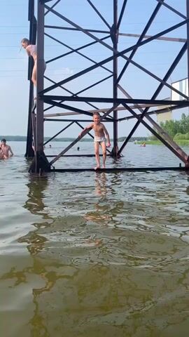 A boy in swim trunks stands on a metal platform of a large industrial structure submerged in water. He looks to his left at another boy who is on the platform above him. The boy on the lower platform then jumps into the water, creating a splash. To the left, a girl in a swimsuit is climbing up the metal structure. Other people are visible in the water further in the background. The sky is blue and clear, with some clouds. The water is murky and reflective, showing the structure and the sky.