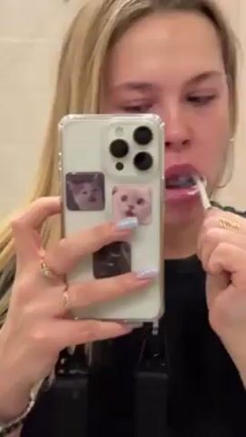 A woman with blonde hair is brushing her teeth in front of a bathroom mirror. She is holding up her phone to record herself. The phone case is transparent, and has three square pictures of cats stuck to it. The bathroom has white tiled walls.
