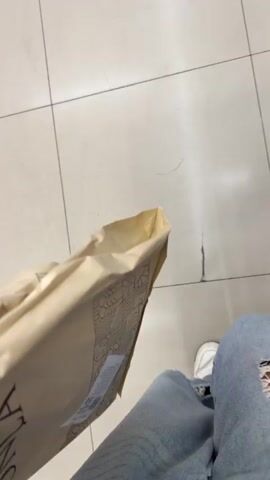 A first-person perspective video shows a person holding a light brown paper bag. The person is wearing ripped denim jeans and white sneakers. The background is a tiled floor with visible grout lines. The camera then pans to show a blurry scene of people and shopping carts. One person has long, dark red hair and is wearing dark pants and a multi-colored shirt. Another person is visible wearing dark pants. Two shopping carts with black baskets and orange accents are also visible. The video continues to pan, showing more of the store environment with blurred shelving and merchandise. The camera then returns to the first-person perspective, showing the person's legs and the paper bag again.