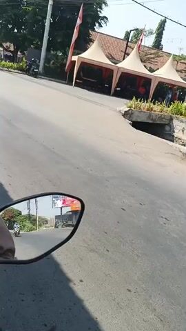 The video shows a view from a motorcycle moving along a paved road. On the right side of the road, there are several tall trees, some green bushes, and a building with a red and yellow striped awning, possibly a convenience store. Flags are seen fluttering in the wind on poles. In the foreground, a motorcycle with a female rider is visible. Further down the road, there are some tents with red coverings, and a few people are seen near them. The sky is clear and bright, indicating a sunny day. The road appears to be in good condition with some cracks visible.