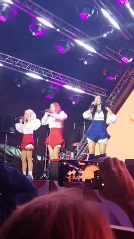 A vertical video shows a live musical performance on a stage. Three women are on stage, dressed in white blouses with puffy sleeves and red skirts or shorts. The woman on the right is singing into a microphone and wearing blue shorts and tall blue boots. The woman in the middle is also singing into a microphone. The woman on the left is also singing. Behind them, a large screen displays images of two women's faces. Stage lighting, consisting of purple spotlights and white strip lights, illuminates the scene. In the foreground, the hands of audience members are visible, one holding a smartphone to record the performance.
