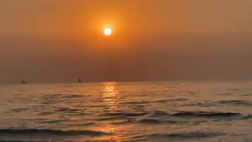 The video depicts a seascape at sunset. The sun, a bright orb, hangs in the hazy, orange-tinted sky, casting a shimmering reflection on the ocean surface. Gentle waves ripple toward the foreground. Two sailboats are visible in the distance, silhouetted against the bright horizon. The overall atmosphere is calm and serene.