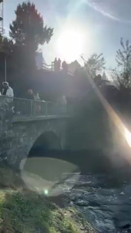 A video shows a flowing river with some rapids.  A stone bridge spans the river. People are standing on the bridge observing the scene.  On the far bank is some forested terrain. The sun is shining brightly.
