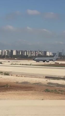 The video shows a large gray military aircraft, possibly a refueling tanker, taxiing on an airport runway. The aircraft has four engines under its wings and appears to be moving slowly. In the background, a dense urban landscape with many tall buildings is visible under a blue sky with scattered clouds. The foreground shows the dusty, undeveloped terrain surrounding the runway, with some sparse vegetation. The lighting suggests it is daytime, likely in the morning or afternoon. The camera is positioned at a distance, giving a wide view of the scene. No people are clearly visible.