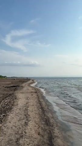 A person walks on a sandy path along the coast. The path is to the left of the video frame, and the ocean is to the right. The sky is blue with scattered clouds. The video appears to be taken during the daytime.