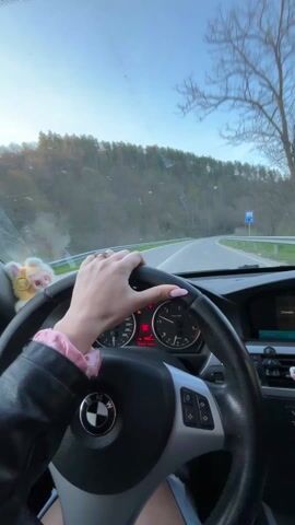 The video shows a first-person view from inside a car driving on a winding road. The driver is a woman with pink nail polish and a ring on her finger, wearing a black leather jacket with a pink scrunchie on her wrist. Her hand is on the steering wheel of what appears to be a BMW. The car's dashboard and infotainment system are visible, with the screen displaying options for climate control, communication, and entertainment. The road is surrounded by trees and some vegetation, and the sky is visible through the windshield. The car is moving at a moderate speed along the road.