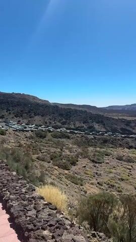 The video pans across a barren, volcanic landscape under a clear blue sky. The terrain is rocky and arid, with sparse, dry vegetation and dark, hardened lava flows visible in the distance. In the mid-ground, a road runs along the edge of a slope, with a long line of parked cars, suggesting a popular tourist destination. Further into the scene, a large, dark building with a red roof sits on a hill. The camera then focuses on a pathway bordering the road, with a stone wall on one side and more vegetation. It then moves to show people walking along the paved path and more cars parked along the roadside. The video ends by showing a closer view of the parked cars, including one silver car with a license plate reading "2685 LYT".