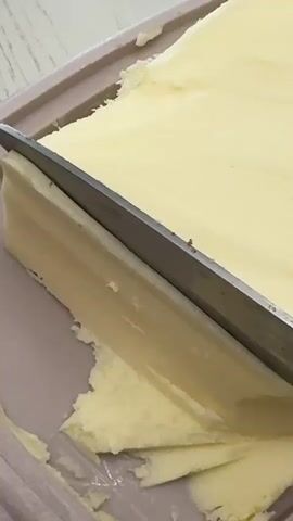 In a close-up indoor shot, a silver knife is shown slicing horizontally into a large, rectangular block of a pale yellow, soft substance, likely butter or margarine. The knife has already cut partway through, separating a thin, uniform layer from the main block. Crumbled pieces of the yellow substance are visible on the light-colored surface surrounding the block, suggesting its soft texture. The surface the block rests on appears to be a light beige or pinkish-brown container or cutting board. The scene takes place in Yuzhny, Russia.