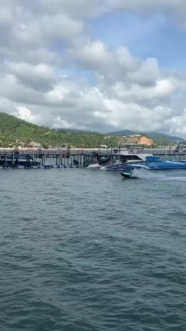A jet ski with a rider makes a sharp turn in the water, creating a spray of water as it moves past a pier. The pier is lined with several other jet skis and some inflatable water toys. In the background, lush green hills rise behind a village. The sky is partly cloudy.