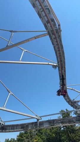 A roller coaster car with two people in it hangs upside down from a track against a clear blue sky. The car is red and black, and the track is made of metal. The roller coaster is on a large, curved structure supported by metal beams. In the background, there are trees and a distant landscape with mountains.
