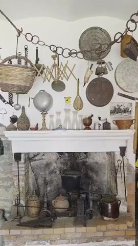 The video pans across a room displaying a collection of antique kitchenware and tools, likely in a historical home or museum. The focus is on a large, stone fireplace and the mantelpiece above it.

On the mantelpiece, an assortment of glass bottles of various sizes and shapes are arranged alongside a mortar and pestle, a spice grinder, and a small bowl. Behind these, a decorative, metallic orb on a stand is visible, as well as a small, brown, ceramic pot. To the left of the mantelpiece, a vintage clock is mounted on the wall.

The fireplace itself contains various cooking implements, including a large black pot hanging over the hearth, a smaller pot on the grate, and several metal tools. A large, woven basket hangs from chains above the mantelpiece.

The walls are adorned with numerous metal tools, including knives, tongs, and what appear to be farming implements. Some of these are arranged in a grid-like pattern. The walls are also decorated with a collection of framed photographs and unframed prints, some of which appear to be old family portraits.

To the left of the fireplace, a large, perforated metal pan hangs on the wall, and below it, a spade and a rectangular wooden tray are propped against the wall. Several large, cylindrical objects, possibly barrels or storage containers, are visible in the lower left corner of the frame. A large, metal milk can also stands near the fireplace. The overall impression is one of a well-preserved historical domestic setting, showcasing the tools and artifacts of a bygone era.