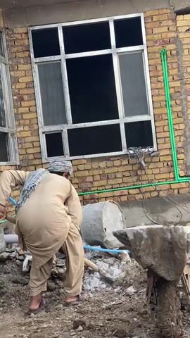 A man in a tan shalwar kameez and a checkered headscarf is shoveling material from the ground into a concrete mixer. The concrete mixer is a large, round, gray drum on its side. The man is facing away from the camera, with his back bent over as he uses a shovel. In the background, a brick building has a large, multi-pane window with a silver frame. Green pipes run vertically along the side of the building near the window. Debris, including rocks and dirt, is scattered on the ground around the man and the mixer. Another person's arm and hand, holding a shovel, is visible on the left side of the frame.