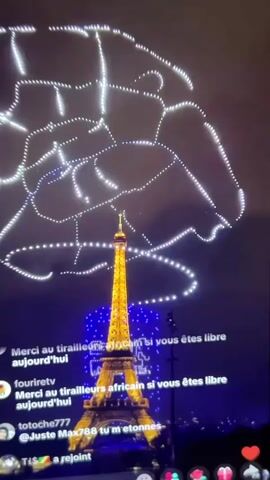 A night view of the Eiffel Tower in Paris is visible. The Eiffel Tower is illuminated with golden lights. Above the tower, a drone show displays patterns of white lights in the night sky. The lower portion of the screen shows a live stream interface with comments from viewers. Some comments express thanks and greetings. The overall scene is dark with the illuminated tower and the light patterns from the drones being the primary visual elements.