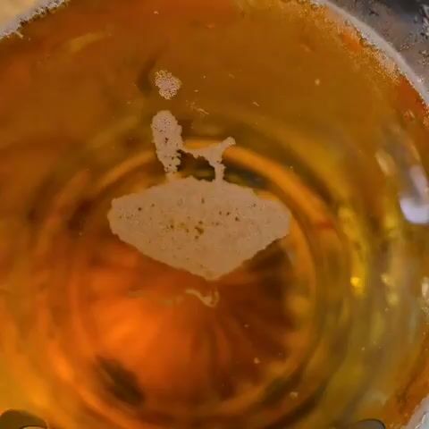 The video shows a close-up of a glass containing a yellow liquid. There is a foamy residue at the top of the liquid, in the shape of a figure sitting in a chair. The background sounds include general crowd noise.
