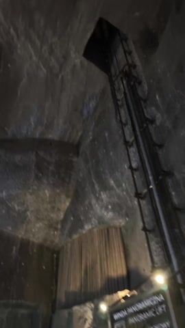 The video shows a view inside a cavernous space with rough, rocky walls. The lighting is dim, with a warm, orange glow illuminating certain areas. There are wooden structures built within the cavern, resembling platforms or walkways. The camera appears to be moving, panning upwards to show the full extent of the interior.