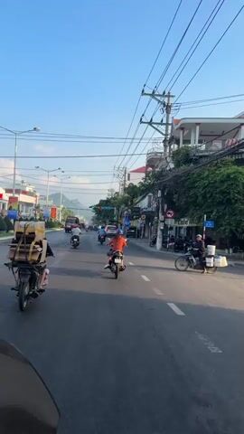 This is a first-person perspective video taken from a motorcycle in a city. The video shows a busy street with several other motorcycles and a few cars. The sky is clear and blue, indicating it is daytime. The buildings lining the street appear to be commercial establishments with signs in a foreign language. The motorcycle in front of the camera is carrying a large stack of cardboard boxes strapped to its rear rack. The rider of this motorcycle is wearing a dark jacket and is partially visible. Another motorcycle with a rider wearing an orange shirt is visible to the right. Utility poles with wires run along the street. The overall impression is of a typical urban scene with a lot of motorcycle traffic.