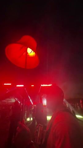 The video shows a crowd of people at an outdoor concert at night, illuminated by red stage lights. A person in the foreground holds up a tall lamp with a red lampshade, making it stand out against the night sky. The stage is visible in the background with lights flashing and moving. People are moving and seemingly enjoying the concert. The scene is dark except for the stage lights.