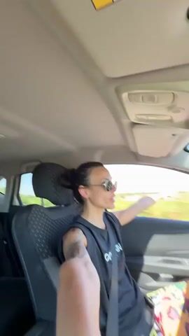 The video shows a woman driving a car on a sunny day. She is wearing sunglasses and a black tank top. She has her right arm extended out the window, and her hand is raised. Her hair is pulled back in a bun. The interior of the car is visible, including the dashboard and seats. The scenery outside the window appears to be rural, with trees and fields. The woman seems to be enjoying herself.