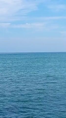 A jet ski speeds across the open ocean, leaving a trail of white spray in its wake. The water is a deep blue, and the sky above is a clear, bright blue with a few scattered white clouds. The sun is shining, and the overall scene is one of a beautiful, sunny day at the beach. The jet ski is moving from left to right in the frame. The rider is wearing a helmet and appears to be enjoying the ride. The camera is focused on the jet ski and the water, and the background is blurred. The video is short and to the point, capturing a brief moment of fun and excitement.