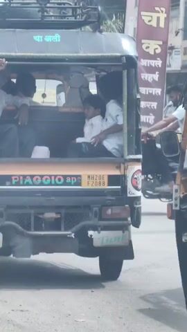 The video shows the rear view of a Piaggio Ape auto rickshaw with several people inside. A man in a light-colored shirt is visible in the driver's seat, waving his hand. Two children are seated in the back passenger area, one wearing a white shirt and the other a blue shirt. The auto rickshaw has a black roof with some text in a foreign language, possibly Marathi, that reads "चा लाली" (Cha Laali). A license plate with the characters "MH20E F2088" is visible on the back. The auto rickshaw is on a paved road, possibly in a city, with other vehicles and buildings in the background. Another auto rickshaw is partially visible on the right side of the frame. The overall scene appears to be a typical street view in India.