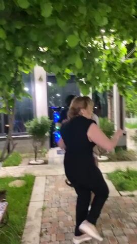 Two women dance to music outside a building. One woman wears a black dress and a head covering, the other wears a black vest and pants. They dance in front of a building entrance, next to trees and greenery. Light is shining from the building and trees are lit up overhead. There is a sign with the number 4 on the building. The ground is made of brick pavers. Other people are visible in the background.