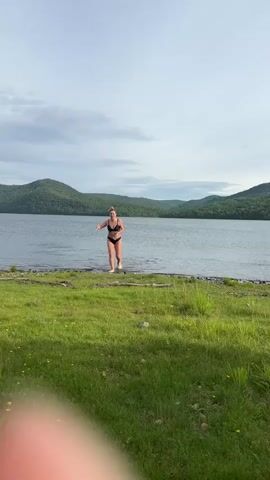 A woman in a black bikini emerges from a lake and walks onto a grassy shore. She has blonde hair pulled back in a bun and is smiling. The lake is surrounded by lush green hills under a cloudy sky. The woman pauses on the shore and adjusts her bikini top.