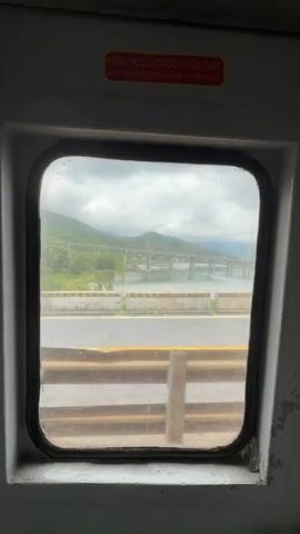 The video shows a view from a vehicle window looking out at a bridge and a body of water. The window is dirty and smudged. Outside the window, a road runs parallel to the water, and a long bridge with many pillars spans the water in the distance. Mountains covered in greenery are visible on the other side of the water. The sky is cloudy. A yellow stripe is visible on the road, and a guardrail runs along the side of the road. The interior of the vehicle is visible in the foreground, including a metal railing and the edge of the window frame. There are no people or animals visible in the video. There is a red sign above the window that appears to be in Vietnamese.