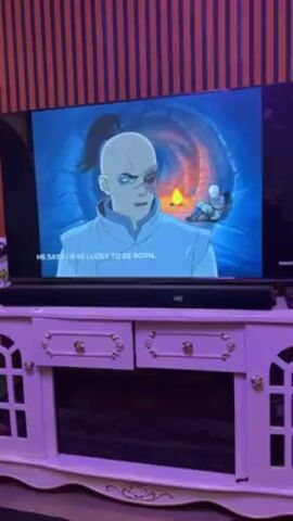 Here's a description of the video:

The video shows a TV screen displaying an animated scene from Avatar: The Last Airbender. On the screen, a character with a ponytail and a scar over his eye is speaking a line about luck. In the next frame, a bald character is shown bound in rope, with the first character standing over him. The TV is set on top of a white and gold entertainment center with drawers and a cabinet beneath. Above the entertainment center is a black soundbar. The room appears to have wooden slat wall decor, and the lighting is primarily purple. The room gives off a cozy or entertainment-focused atmosphere.