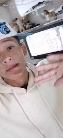 A young man wearing a white baseball cap with a blue design and a tan hoodie looks directly at the camera. He sticks out his tongue and gives a thumbs up.  Behind him, there is a shelf with stuffed animals, a picture frame, and other decorative items.