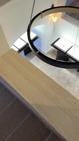 From an elevated vantage point, the camera pans slowly across the interior of what appears to be a jewelry store or gallery. The space features a neutral color palette, primarily whites, beiges, and blacks. The flooring is a light gray carpet with inlaid gold semi-circles forming a pattern.

At the center of the frame is a large, circular chandelier with a black exterior and gold interior trim, suspended from the ceiling by black cables. Below the chandelier are several display tables, each with a black base and a white top showcasing various jewelry pieces.

The walls are painted white, with arched windows or alcoves providing glimpses of staircases and architectural details. The lighting is a mix of ambient and focused, highlighting the jewelry displays. The absence of people contributes to the sterile and pristine atmosphere of the space.