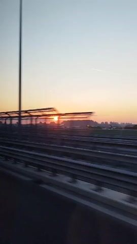 The video captures a scenic view from a moving vehicle on a highway during sunrise. The sun is visible on the horizon, casting a warm glow across the sky. Trees and some distant buildings are silhouetted against the colorful sky, creating a picturesque landscape. The highway guardrail is prominent in the foreground, indicating the perspective from a car. The time of day is clearly early morning, suggested by the sunrise and the soft light. The weather appears to be clear, with a few wisps of clouds and a faint contrail in the sky. The location is Dârvari, Romania, as indicated by the text. There are no people visible in the video, but the presence of the highway suggests human activity and travel. The overall mood is peaceful and tranquil, evoking a sense of journey and the beauty of nature.