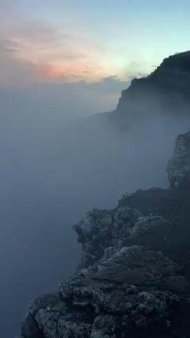 A drone shot pans across a misty mountain landscape. Jagged, dark rocks are visible in the foreground on the right. In the distance, the soft glow of sunrise or sunset is visible through the haze, painting the clouds in hues of pink and orange. The overall atmosphere is serene and ethereal.