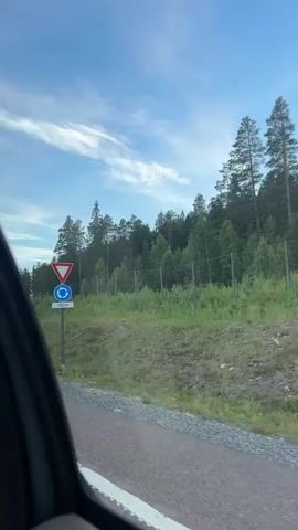 The video shows a view from inside a vehicle traveling along a road. On the right side of the road, there is a traffic sign indicating a roundabout at 150 meters, preceded by a give way sign. Beyond the road is a grassy embankment leading up to a forest. A fence runs along the edge of the forest. Large rocks are visible on the embankment. The sky is blue with scattered clouds. A streetlight is also visible on the left side of the road. The camera pans slightly to the right as the vehicle moves forward.