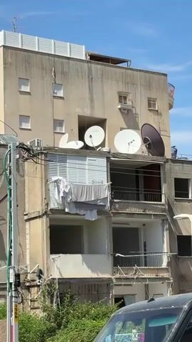 The video presents a static, bright daytime view of a multi-story apartment building in Petah Tikva, Israel, under a clear blue sky. The building's exterior is of a light, possibly beige, color, and it appears somewhat aged.

A notable feature is the collection of four large satellite dishes mounted on a lower rooftop or a prominent balcony level. Three of these dishes are white, while one is a darker shade, possibly black or dark grey. Below this, a balcony on a mid-level floor features white window shutters, with laundry, including white and grey garments, hanging over them to dry. Several other balconies with metal railings are visible on the lower and right sections of the building.

To the left, a utility pole is visible, densely covered with numerous cables and wires that also extend across parts of the building's facade. Small, rectangular windows are spaced across the upper portion of the building.

At the base of the building on the left side, lush green foliage is present. In the bottom right corner of the frame, the front section of a dark-colored car is visible. Text on the car includes the Hebrew word "קפה" (coffee) and the letter "X." The overall scene depicts a typical urban residential environment with strong sunlight. No people or specific activities are observed.