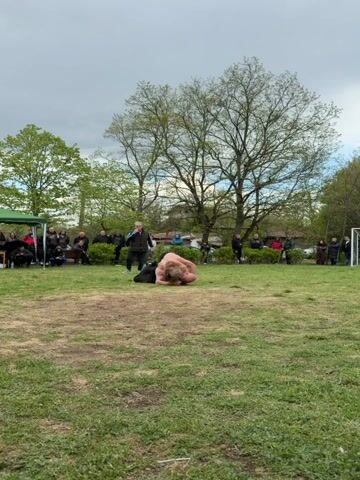 A man lies prone on the grass in a park, stretching his arms and legs, bending and twisting at the waist. The man has bare chest and back, long blonde hair, and wears black trousers and shoes. A man walks toward the prone man. There are people standing and sitting on benches surrounding the field, as well as trees. The sky is overcast.